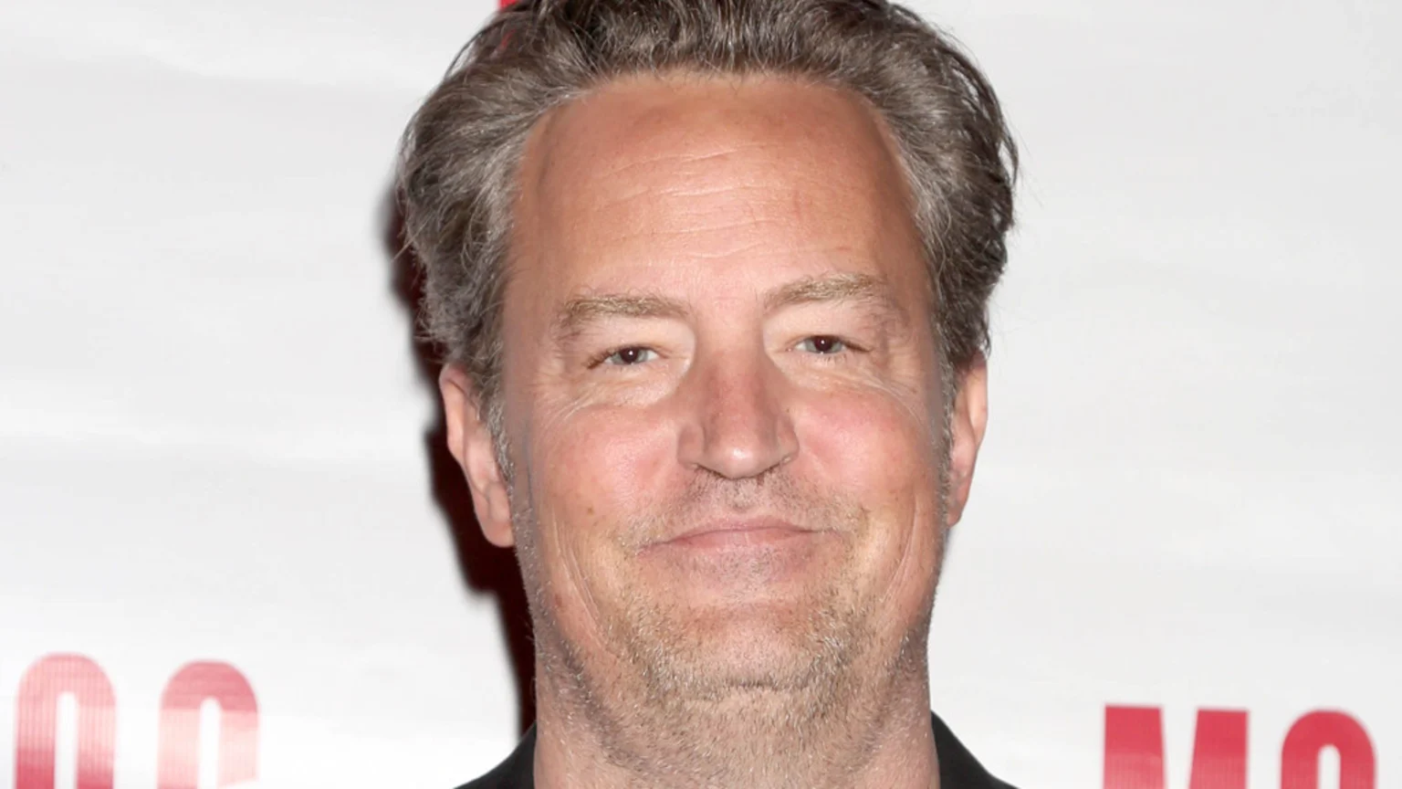 Close-up of Matthew Perry, a man with grey-brown hair and light stubble, smiling subtly directly at the viewer against a light background.