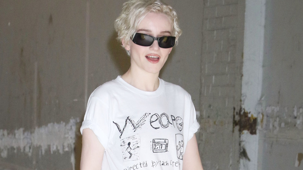 A woman with short blonde hair and sunglasses wears a white graphic t-shirt, smiling slightly with her mouth open against a textured grey wall background.