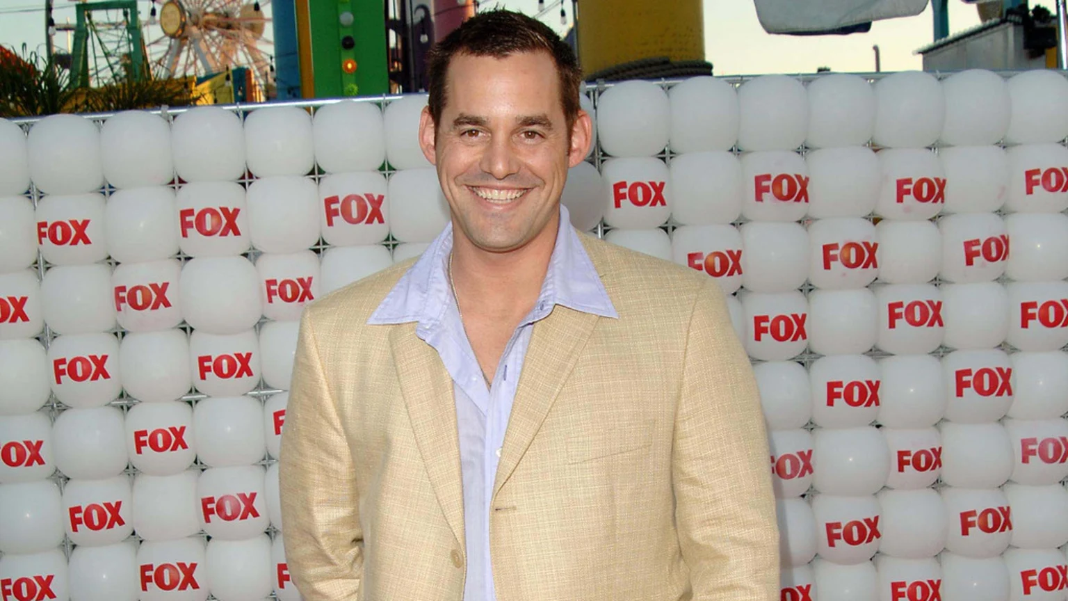 A smiling man in a tan blazer and light blue shirt stands before a wall of white orbs marked "FOX," with amusement park rides visible in the distant background.