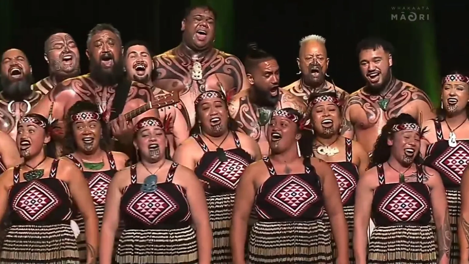 A large group of Māori men and women in traditional attire, many with tā moko, are performing kapa haka, singing with open mouths.