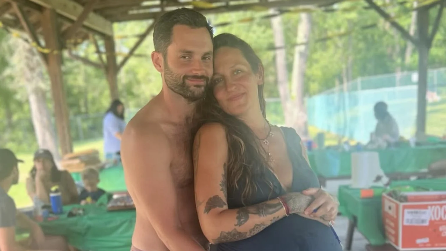 A smiling, bearded, shirtless man embraces a pregnant woman with tattooed arms, both looking at the camera in an outdoor setting with blurred people and tables in the background.