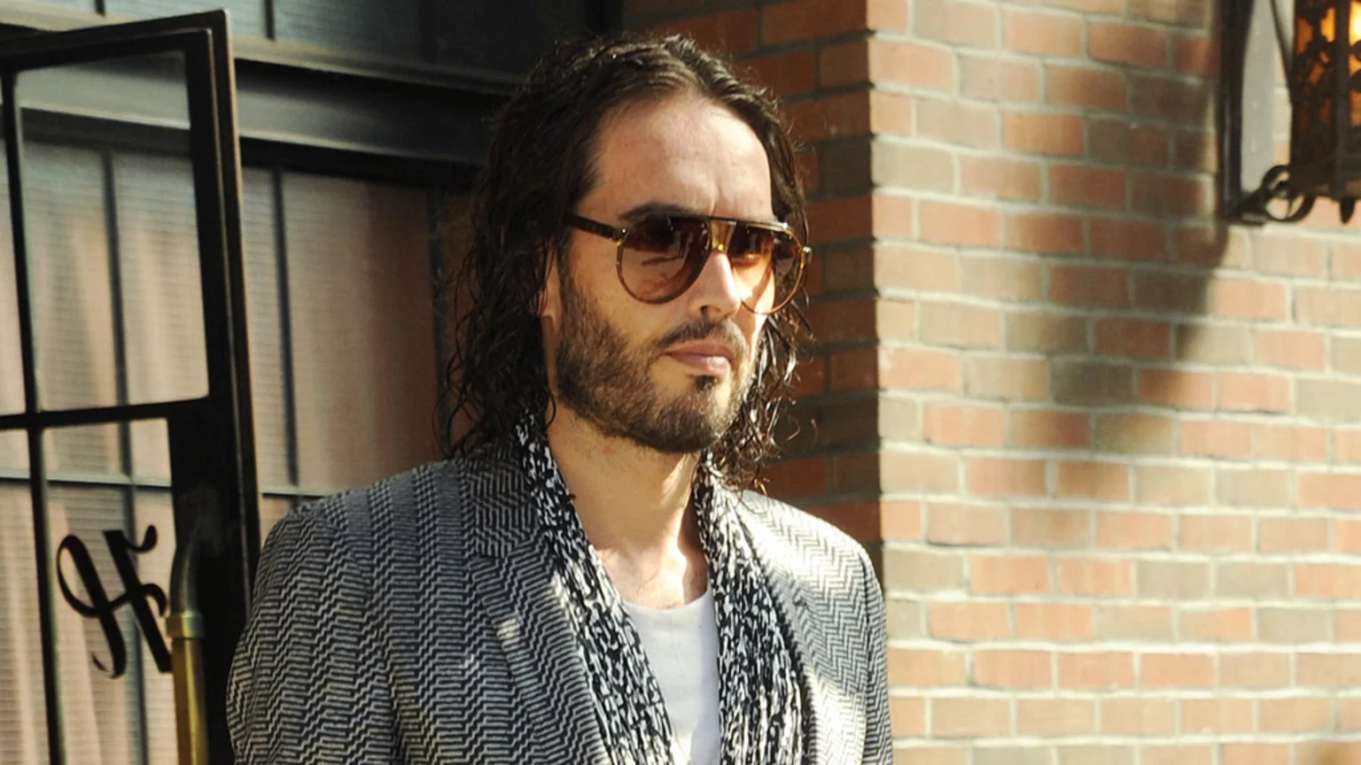 Russell Brand in brown sunglasses, a herringbone jacket, and patterned scarf, looking right against a brick wall and a window with an 'H' logo.