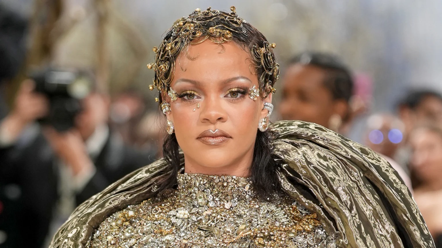 Rihanna, adorned in an ornate gold, jeweled outfit with an elaborate headpiece and face gems, looks directly at the camera at an event.