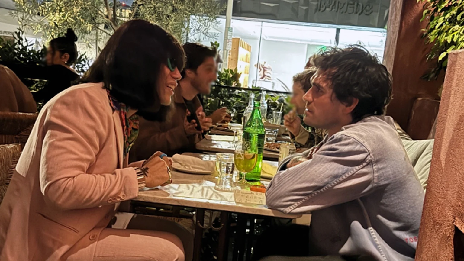Two people, one in a pink blazer smiling at another in a grey shirt, sit at a restaurant table with drinks and food, with other blurred diners and foliage in the background.