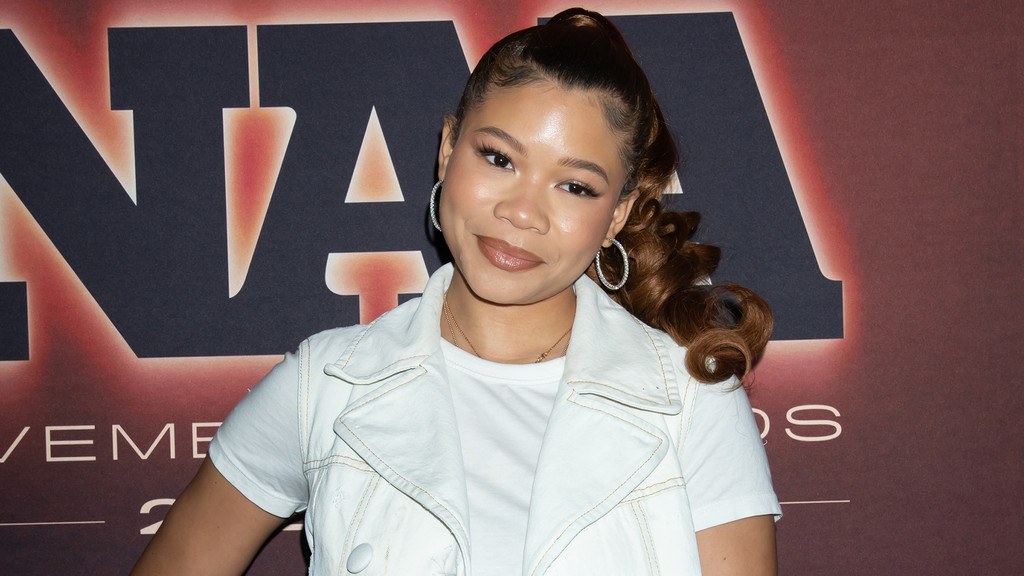 Smiling Storm Reid with brown hair in a high ponytail, wearing a white sleeveless jacket over a white t-shirt, stands in front of a banner with large dark letters.