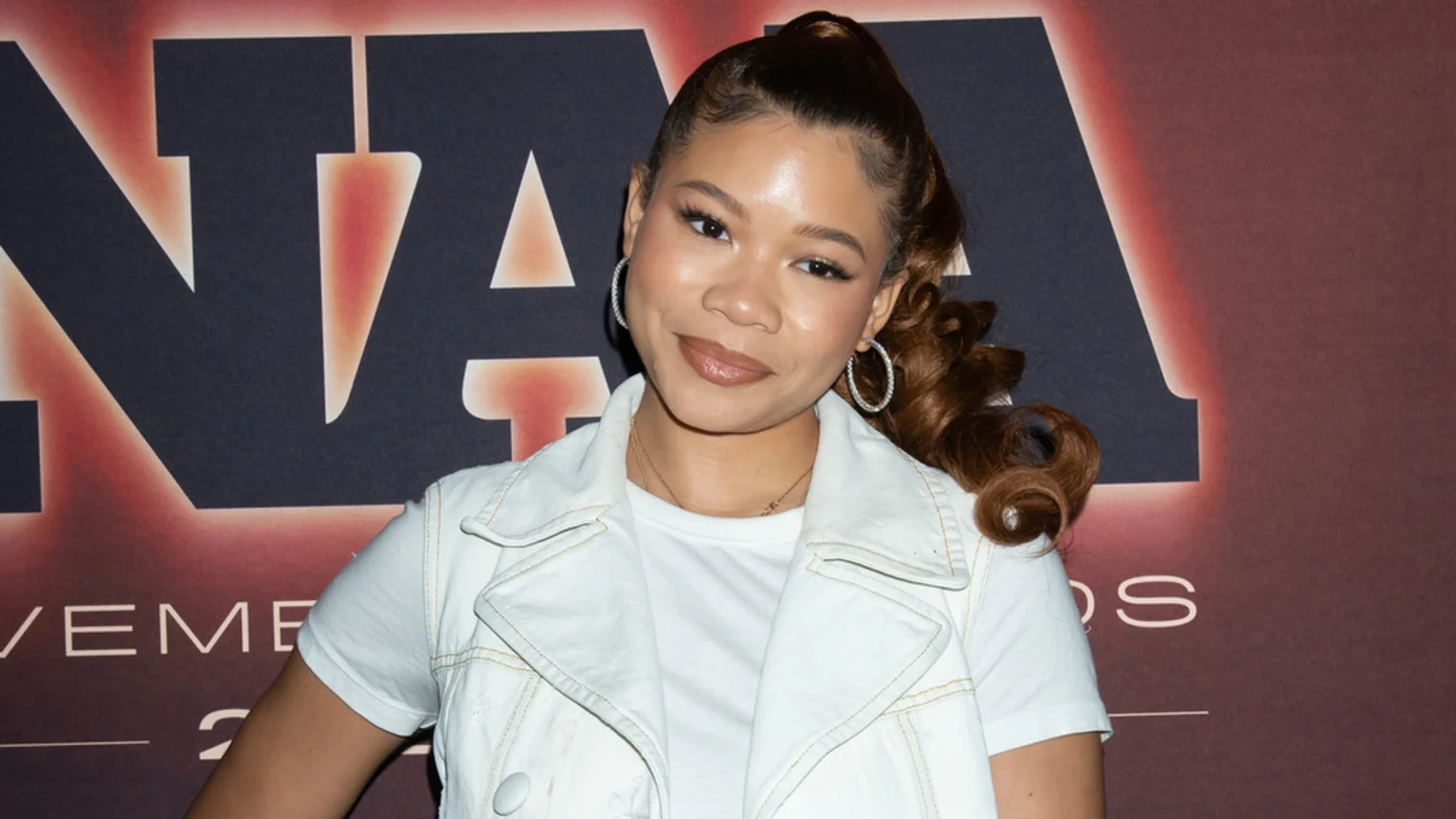 Smiling Storm Reid with brown hair in a high ponytail, wearing a white sleeveless jacket over a white t-shirt, stands in front of a banner with large dark letters.