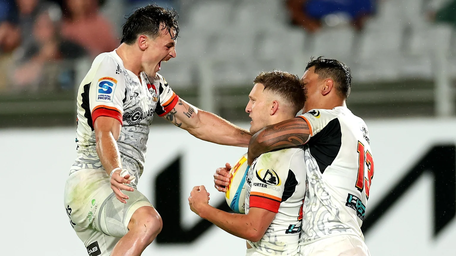 Three Super Rugby Pacific players in white and orange jerseys celebrate excitedly on the field, with one player shouting, another holding a rugby ball, and a third embracing them.