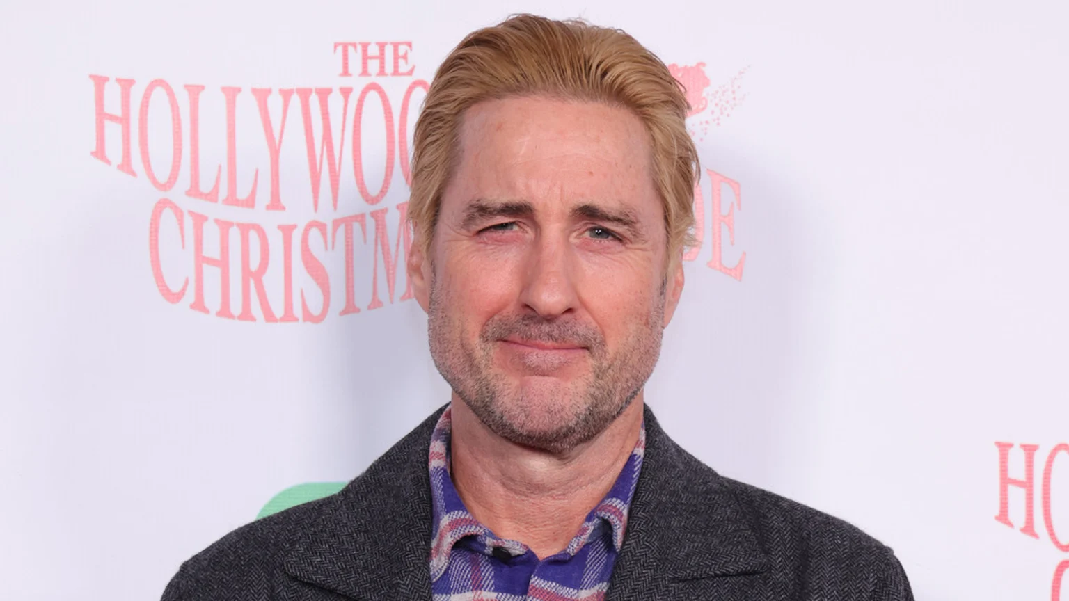 Luke Wilson, with blonde hair and a beard, smiles slightly wearing a dark gray tweed jacket and a blue and red plaid shirt, against a white background with pink "THE HOLLYWOOD CHRISTMAS" text.