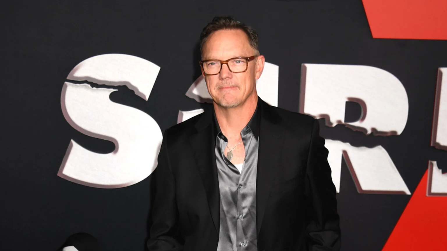 Actor Matthew Lillard, in glasses and a black suit jacket over a grey satin shirt, stands against a dark background with a large, cracked white "Scream" logo.