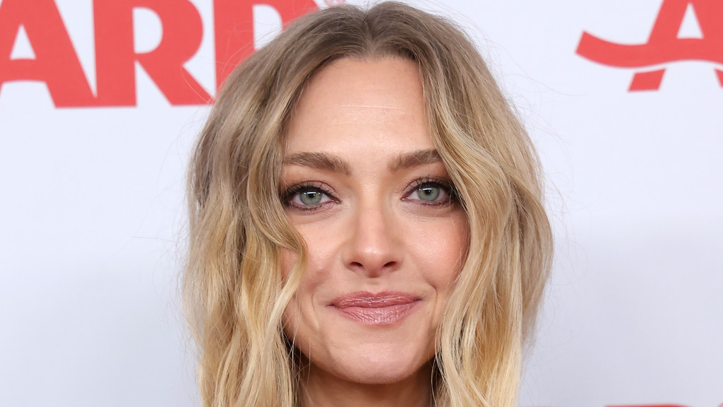 A close-up headshot of a woman with blonde, wavy hair, light green eyes, and a soft smile, against a blurred white background with red letters.