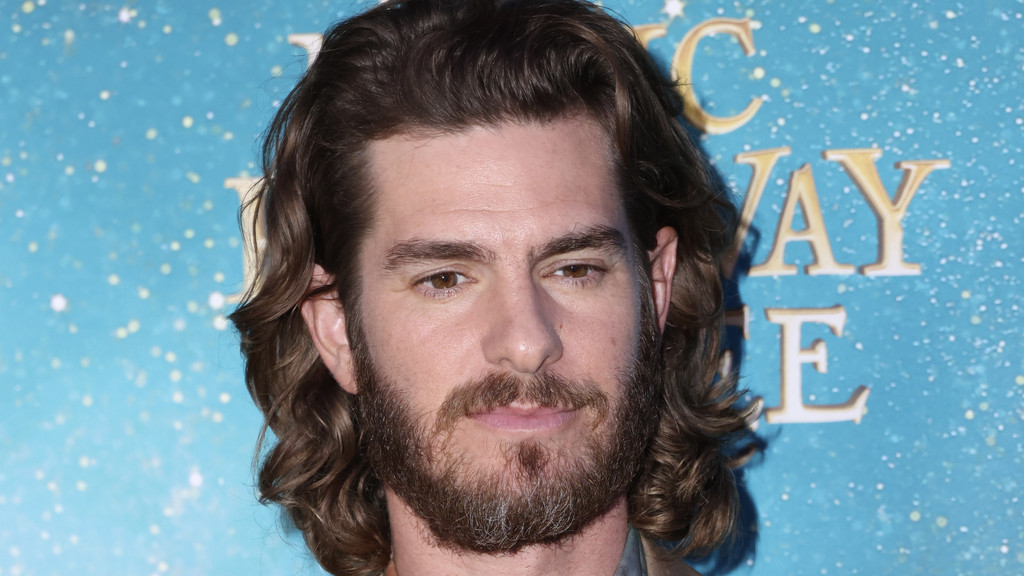 Andrew Garfield, with long brown wavy hair and a full beard, looks thoughtfully against a sparkling blue background with partial gold letters.