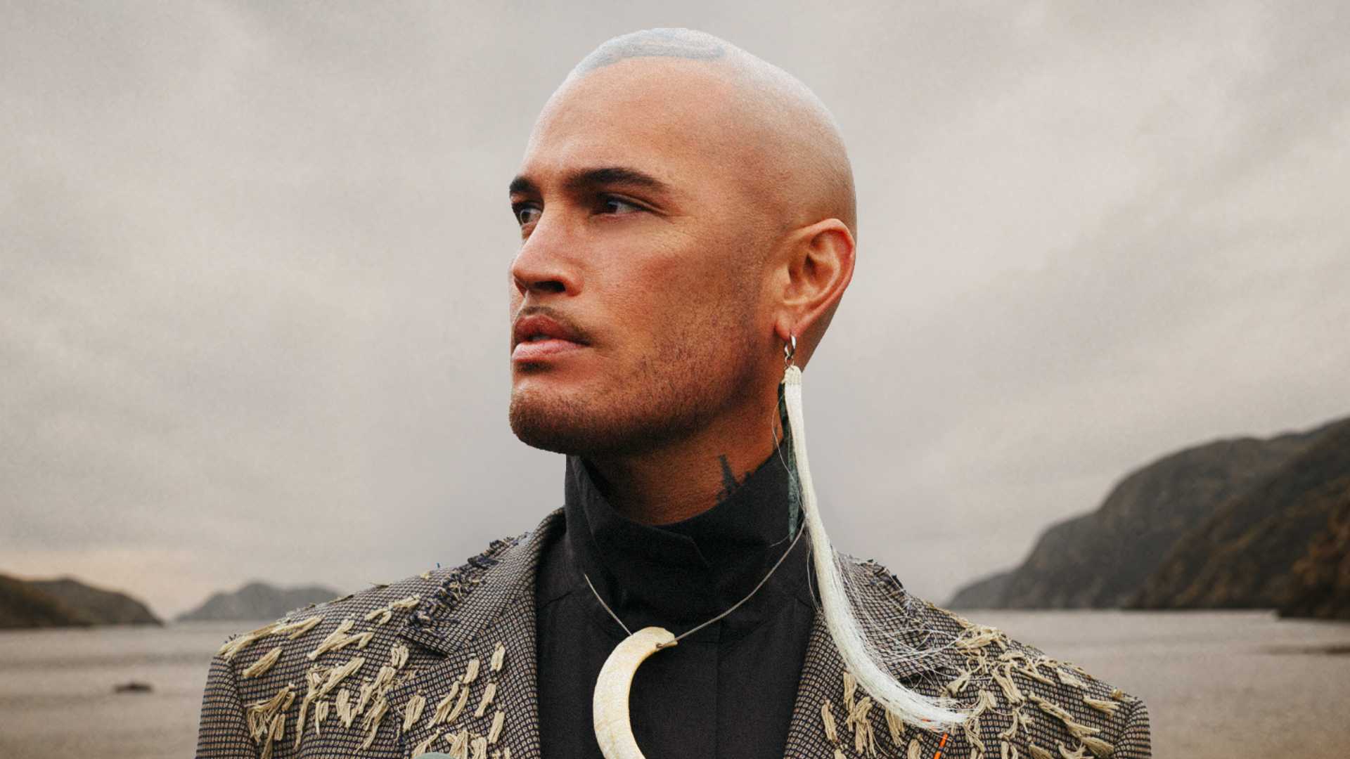 A man with a shaved head, facial hair, a patterned jacket, a long white earring, and a large curved necklace looks left against an overcast landscape background of water and hills.