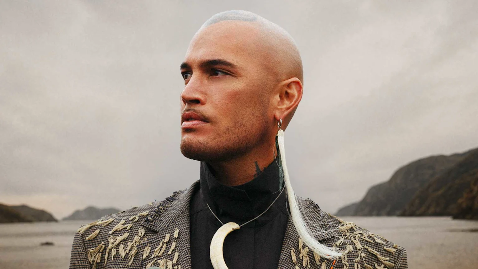 A man with a shaved head, facial hair, a patterned jacket, a long white earring, and a large curved necklace looks left against an overcast landscape background of water and hills.