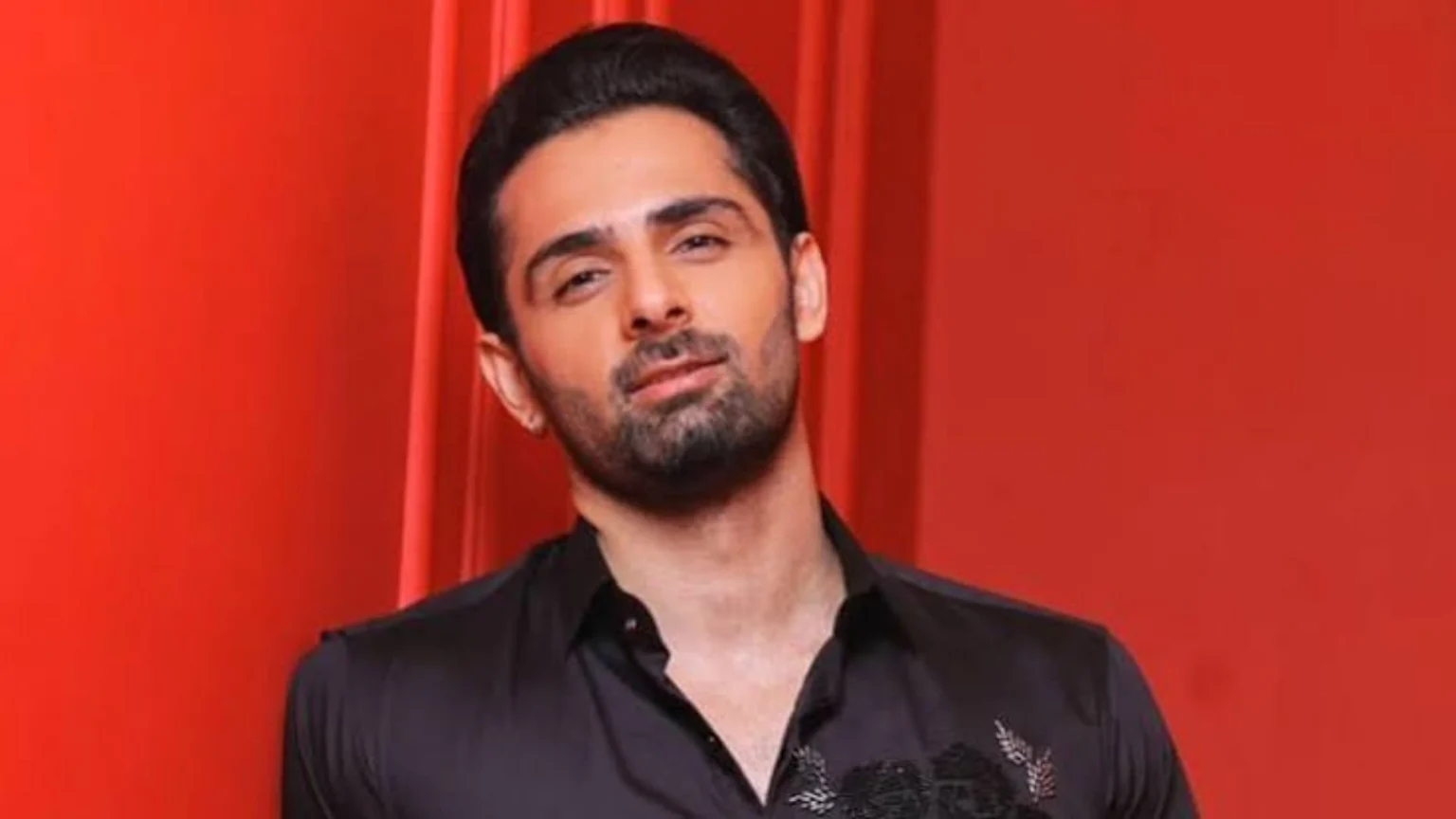 A man with dark hair and a beard, wearing a black shirt with embroidery, looks at the camera against a red background.
