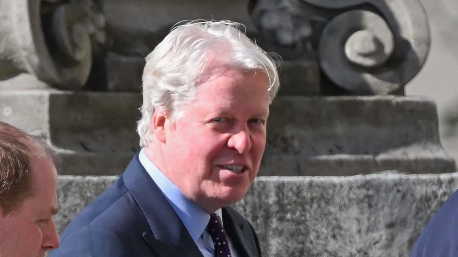 A smiling man with white hair in a dark suit, light blue shirt, and polka dot tie looks at the camera, with blurred stone architecture in the background.