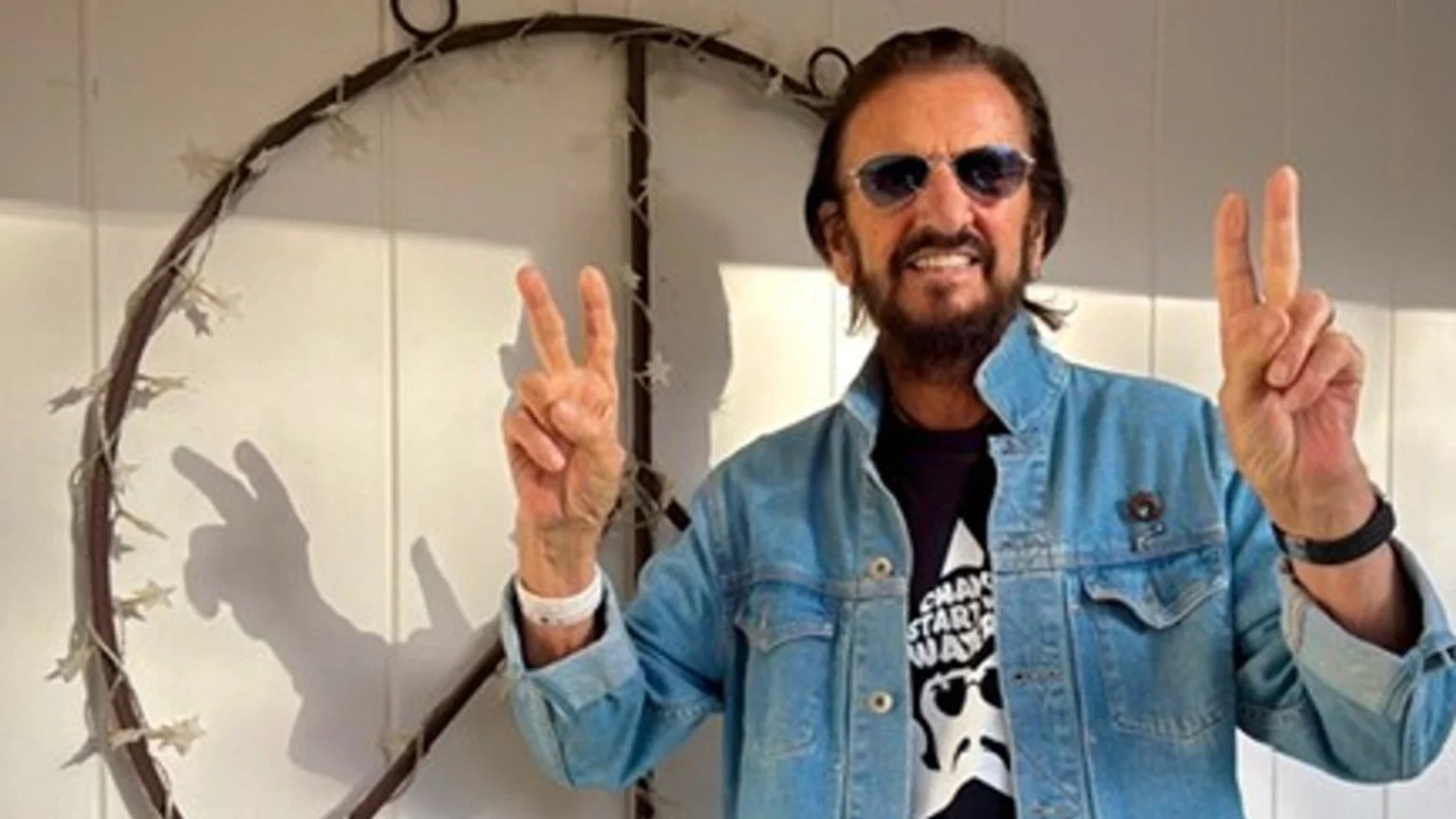 Ringo Starr, smiling and wearing sunglasses and a denim jacket, holds up both hands in peace signs, standing in front of a decorative peace symbol on a white wall.