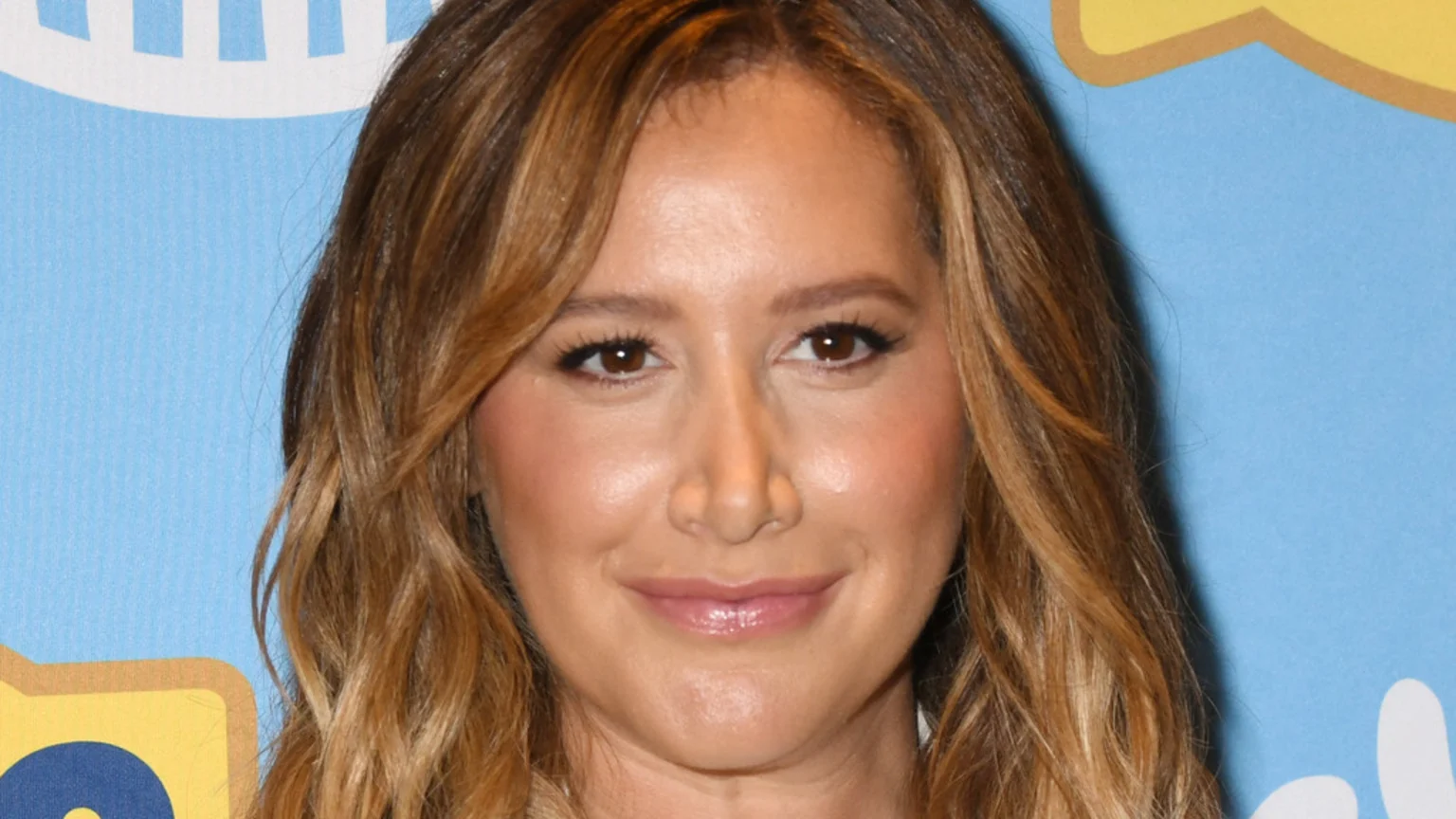 Close-up of actress Ashley Tisdale with long, wavy brown hair, light makeup, and a soft smile, against a light blue background.