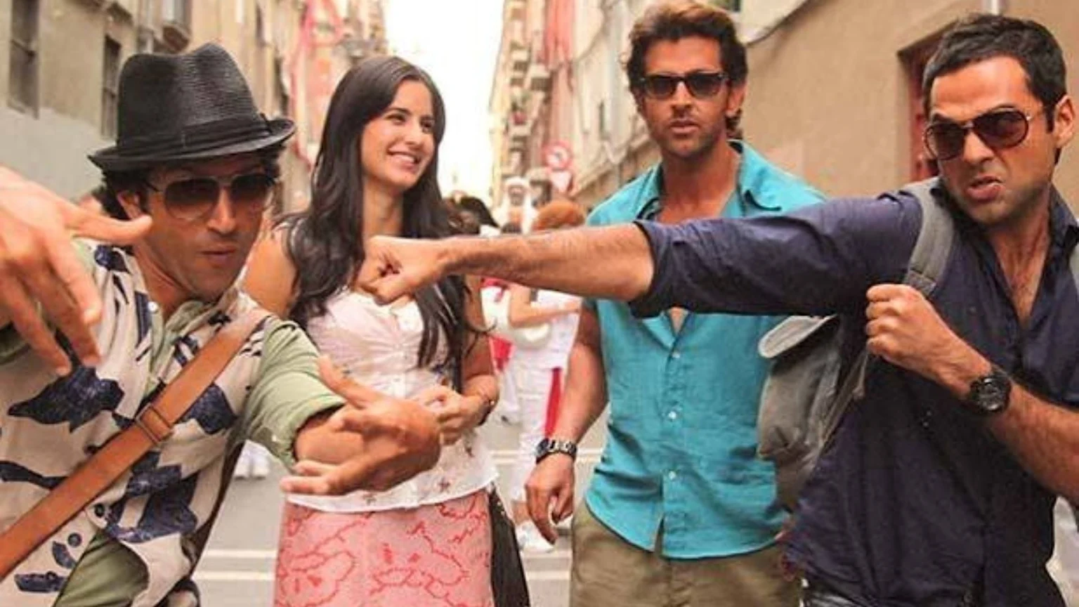 Four people, three men and one woman, pose dynamically on a street, with one man in a hat gesturing, another lunging forward with a raised fist, and the woman smiling between them.