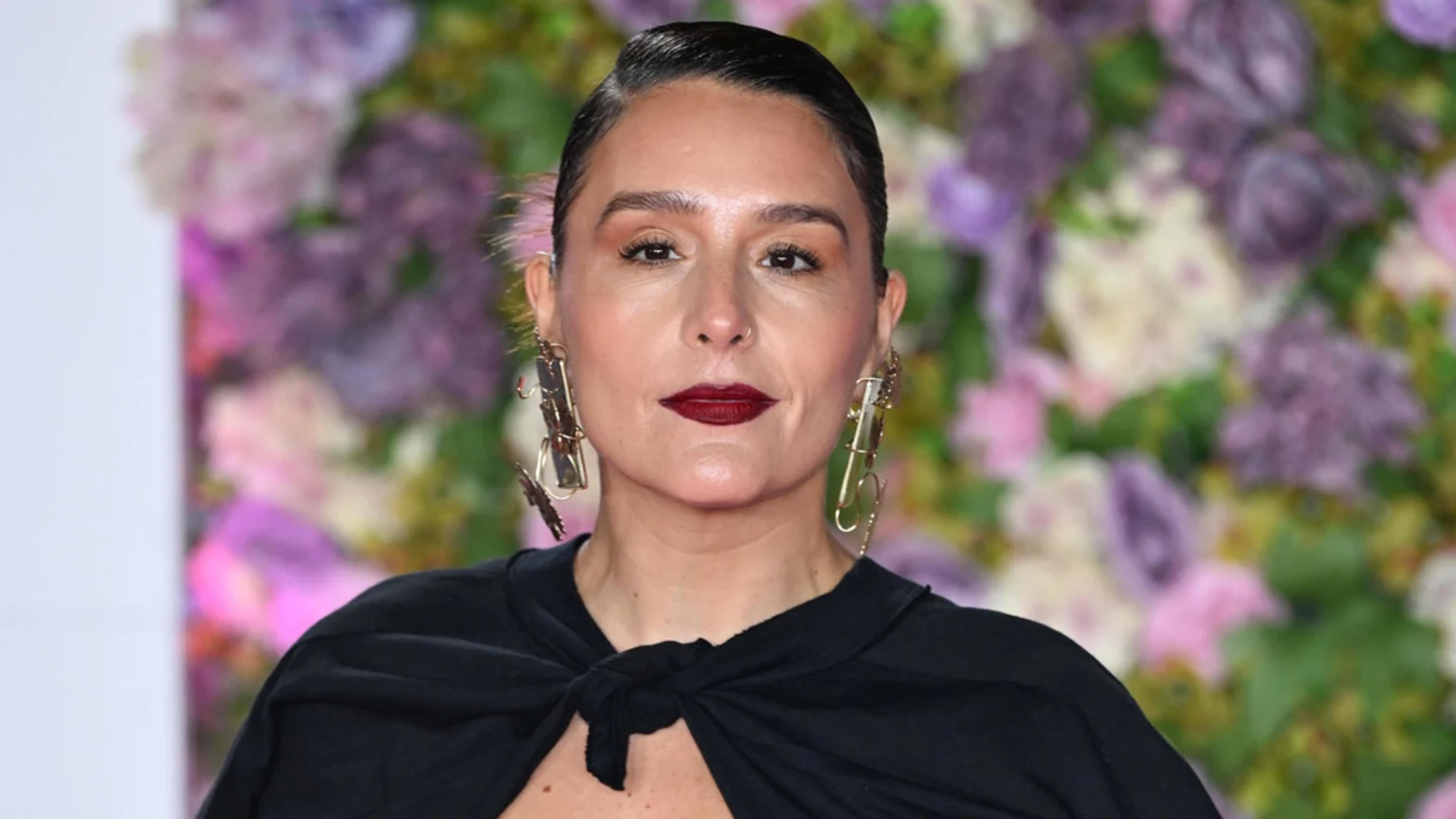 A close-up portrait of an unknown woman aged 30-45, with dark hair pulled back, wearing a black top or cape tied in a front knot. She has large gold-toned dangle earrings, a small stud nose piercing on her right nostril, and dark red lipstick. The background is a blurred floral arrangement in purple, pink, white, and green. The lighting is bright and even, suggesting an indoor event or studio setting.