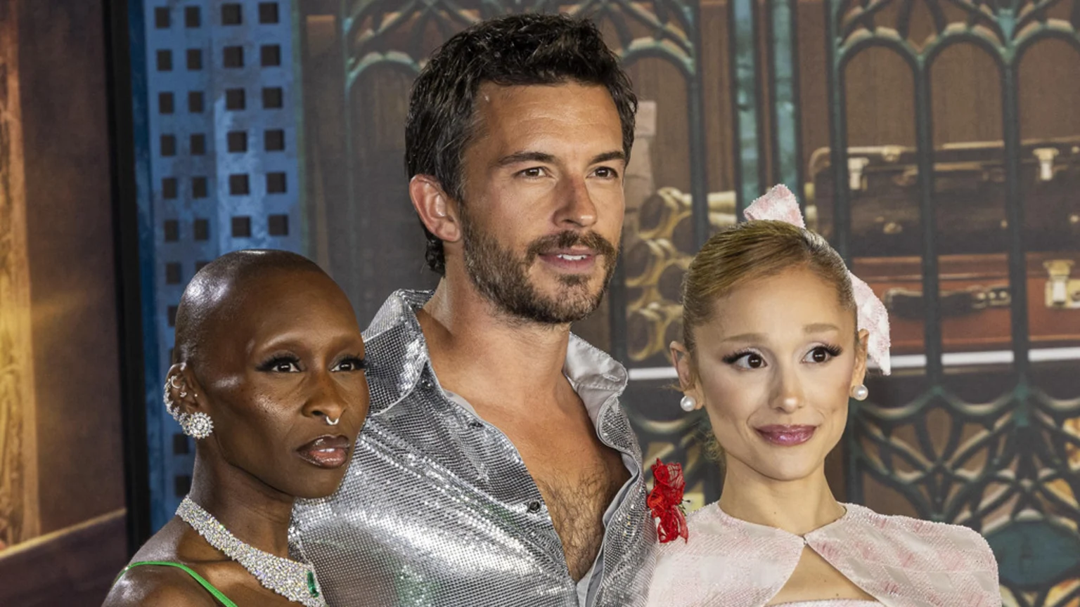 Ariana Grande, Jonathan Bailey, and Cynthia Erivo pose in front of a themed backdrop; Grande wears pink, Bailey a silver shirt, and Erivo a green top.