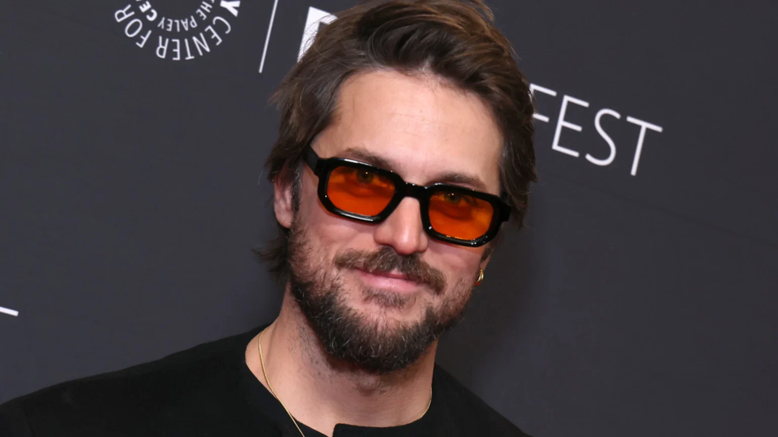 A man with brown hair and a beard, wearing a black shirt, a gold chain, and black-framed sunglasses with orange lenses, smiles slightly in front of a dark gray background with white text.