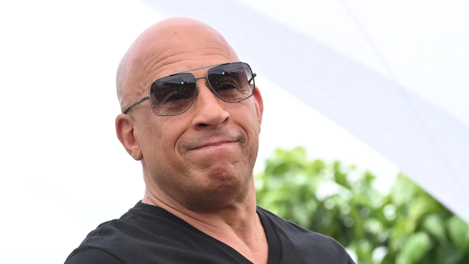 Close-up of actor Vin Diesel outdoors, bald, wearing aviator sunglasses and a black shirt, looking up with a subtle smile.