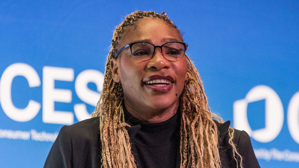A woman with light brown braided hair and glasses smiles, wearing a dark turtleneck and blazer, against a blurred blue background with white letters "CES".