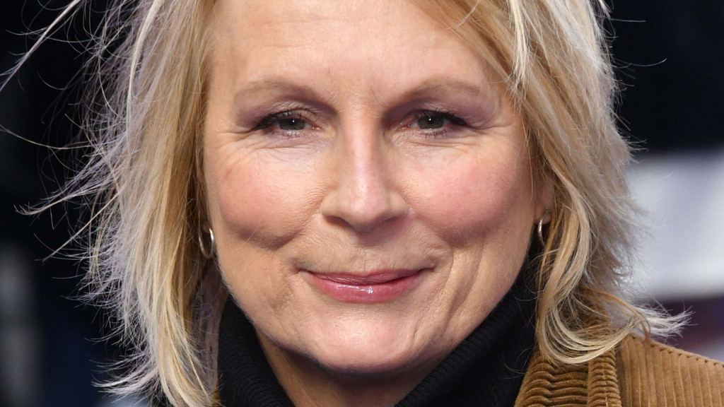 Close-up head-and-shoulders portrait of Jennifer Saunders, smiling gently, with short blonde hair, light makeup, and silver hoop earrings. She wears a black turtleneck and brown corduroy jacket against a softly blurred indoor studio background.