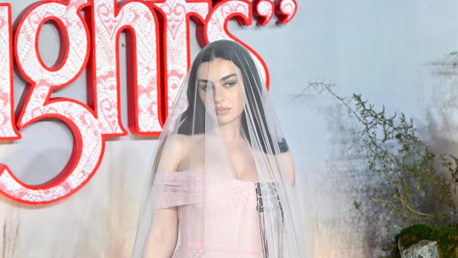 Charli XCX in a pink dress and sheer white veil looks at the camera, with large red and white decorative letters and foliage in the background.