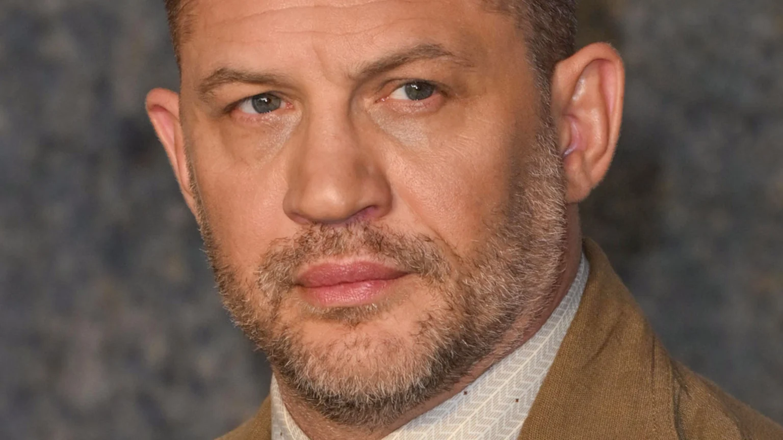 A close-up shot of actor Tom Hardy with a beard, looking directly forward, wearing a patterned shirt and a brown jacket.