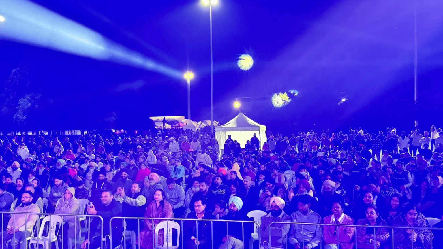 A large outdoor night event with a diverse crowd of people seated and standing, illuminated by blue spotlights and streetlights, with a white tent in the background.