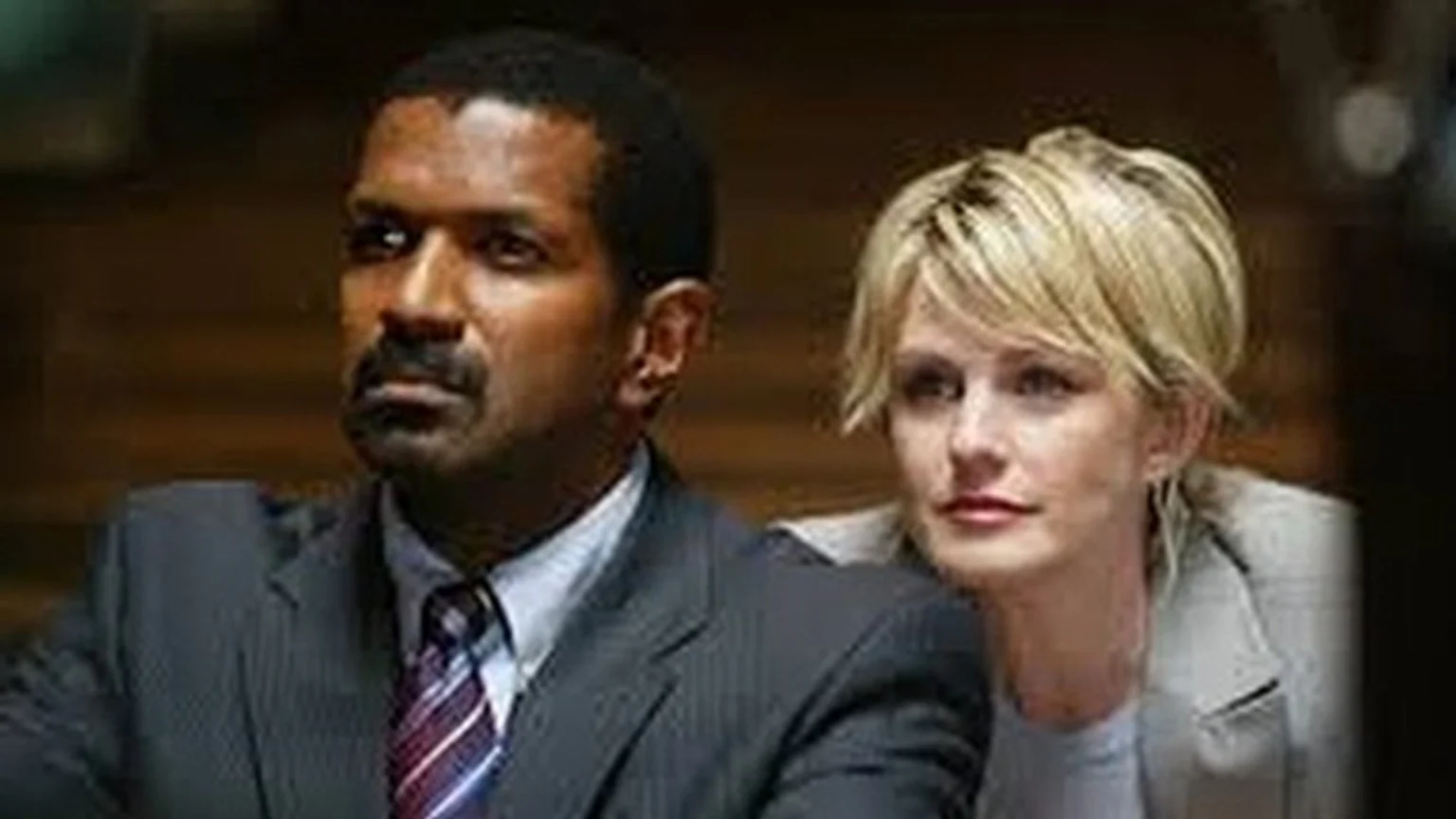 A man with dark skin, a mustache, and a suit looks left, while a blonde woman behind him looks slightly left.