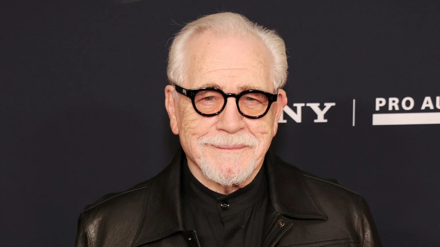 An older man, actor Brian Cox, with white hair and a white goatee, wears black round glasses, a black collared shirt, and a black leather jacket while smiling faintly against a dark background with "SONY" and "PRO AU" logos.