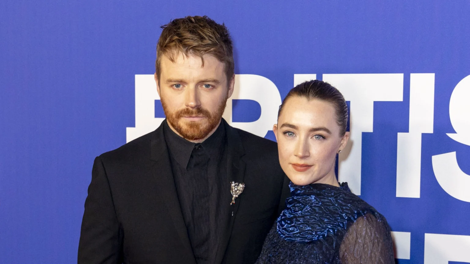 Saoirse Ronan and Jack Lowden pose together, he in a black suit with a brooch, she in a dark blue textured dress, against a blue background with white letters.