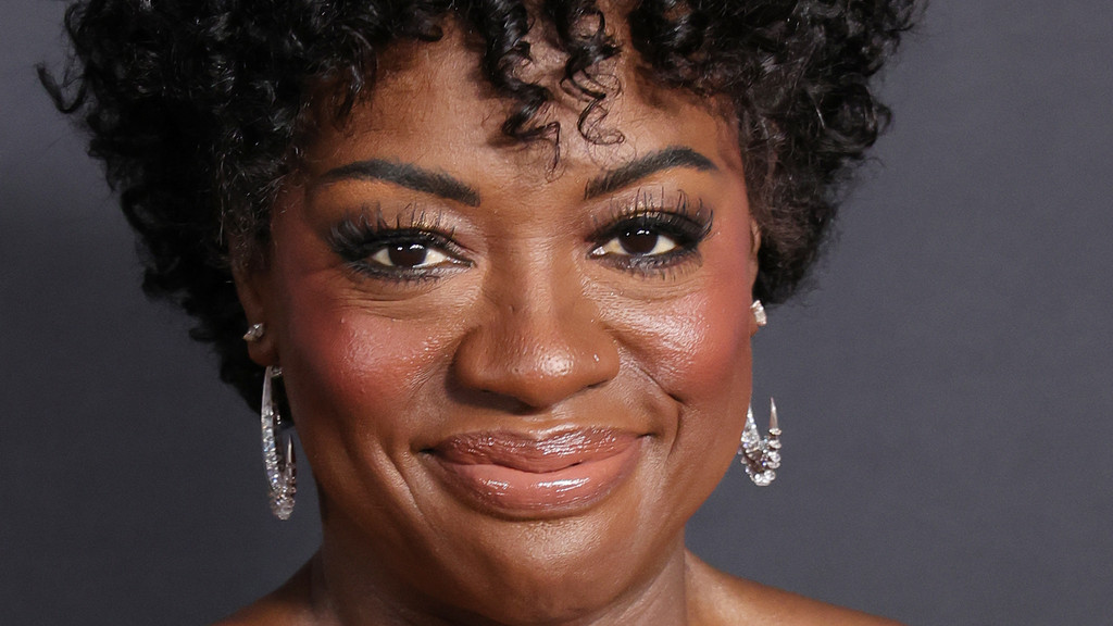 A close-up headshot of Viola Davis smiling warmly, wearing large silver-toned hoop earrings with sparkling stones, subtle makeup, and a formal strapless top. The portrait is set against a plain dark gray studio background with bright, even artificial lighting.