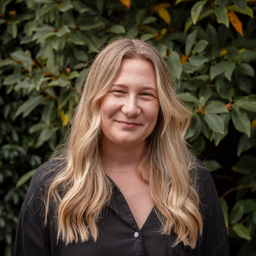 A woman with long blonde hair and a black shirt smiles slightly at the camera against a blurred green foliage background.