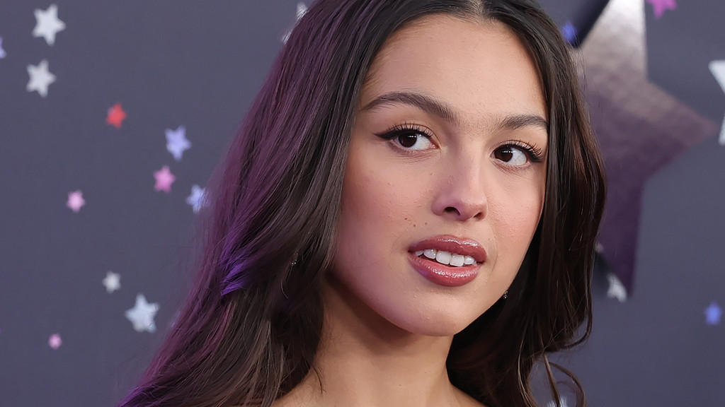 A close-up portrait of Olivia Rodrigo smiling, with long dark hair, winged eyeliner, and glossy lips, set against a dark, blurred background with colorful bokeh stars.