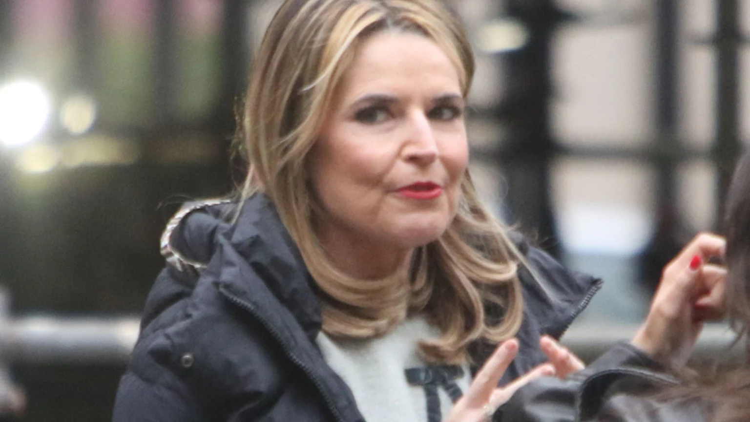 A woman with blonde hair and red lipstick wears a dark puffer jacket over a light sweater with a black bow detail, looking slightly to her right, with a hand with red nail polish visible on the right.