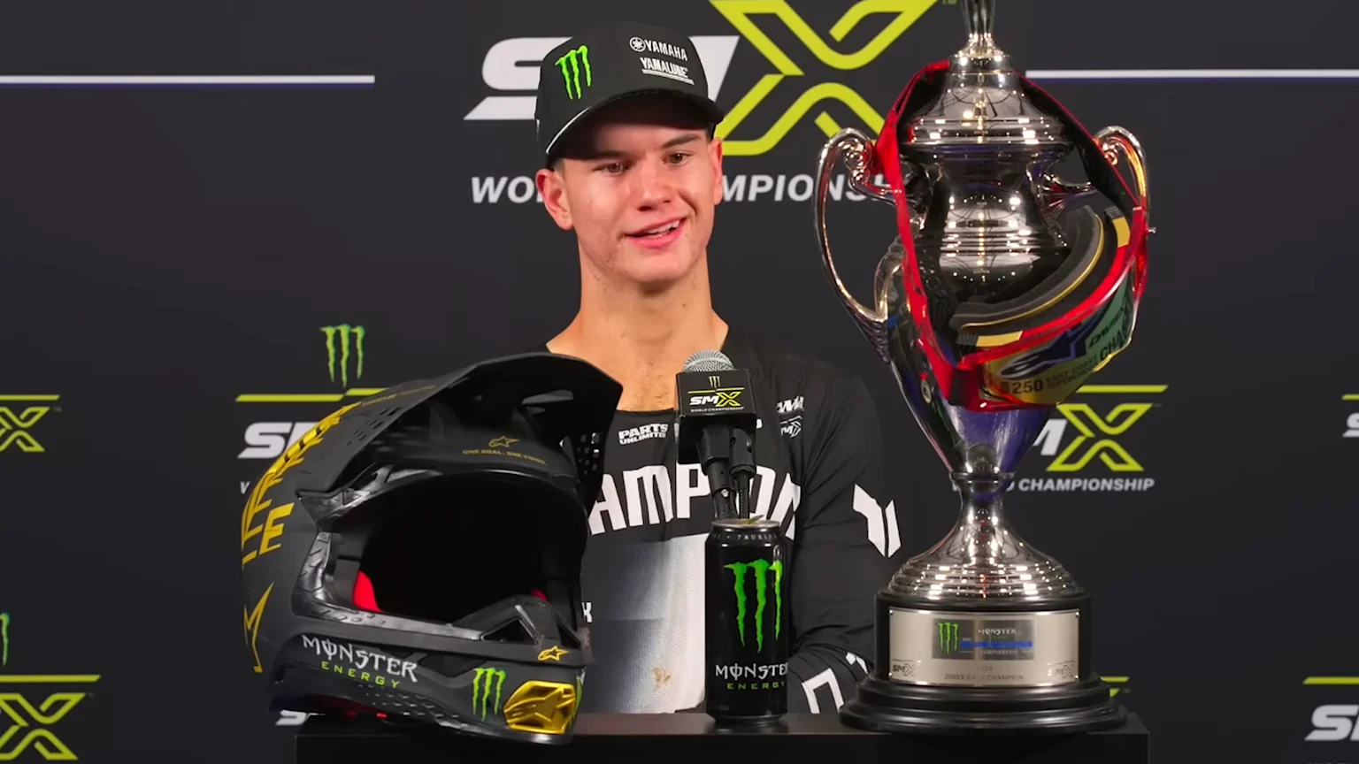 A smiling young man in a cap stands with a championship trophy, helmet, and Monster Energy drink, celebrating his 250SX East Championship win against a black SMX World Championship background.