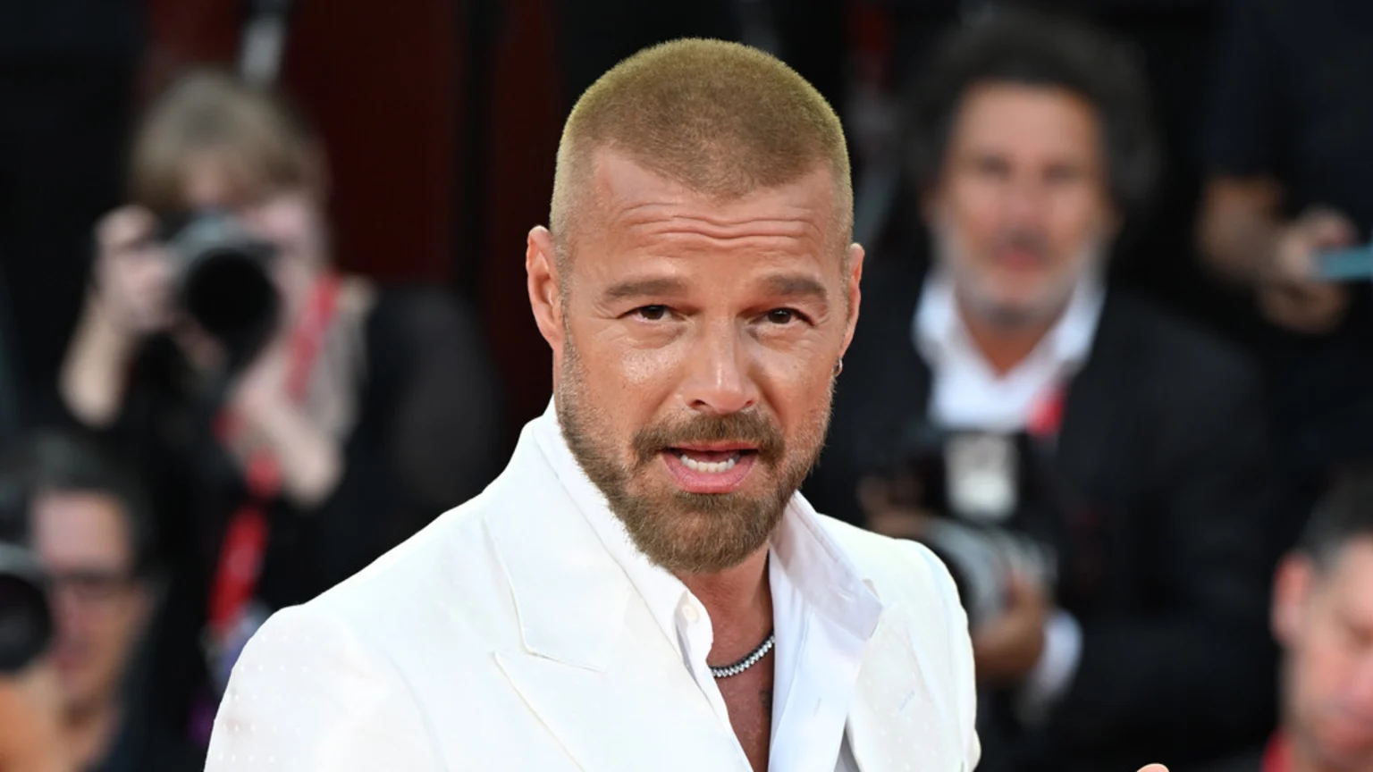 Ricky Martin, with a light buzz cut and beard, wears a white suit and beaded necklace, looking at the camera at a blurred public event.