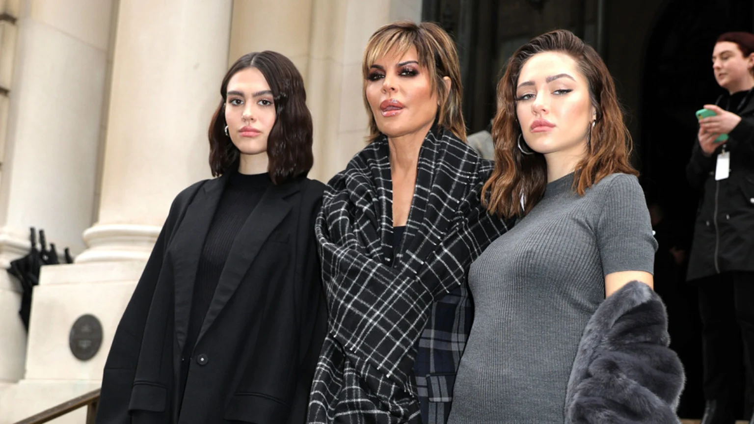Lisa Rinna, wearing a plaid scarf and distinctive makeup, stands between her two daughters; one wears a black blazer, the other a grey ribbed top and holds a fur coat.