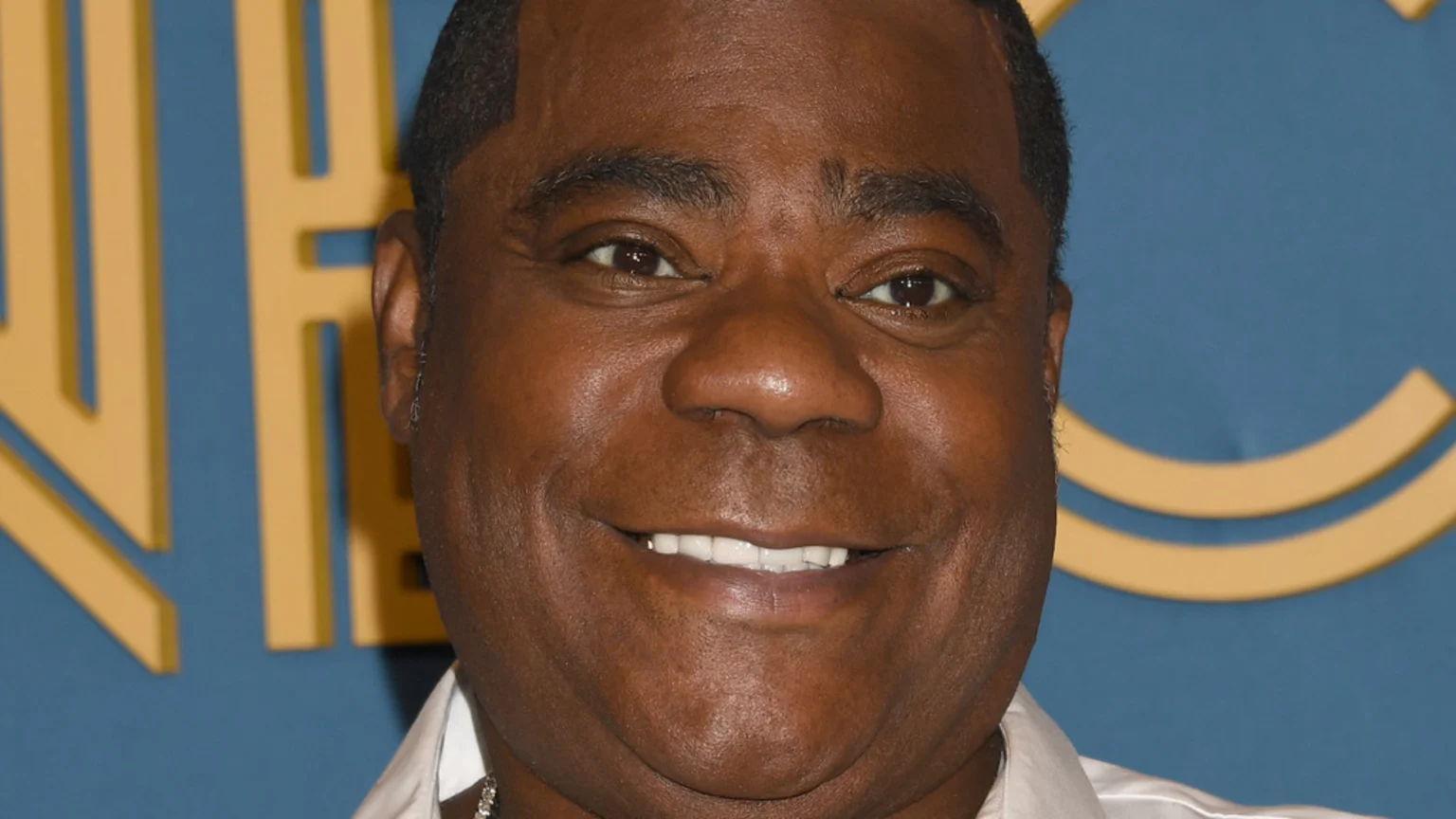 Actor Tracy Morgan smiles broadly, looking directly at the viewer, against a blue background with blurred gold shapes.