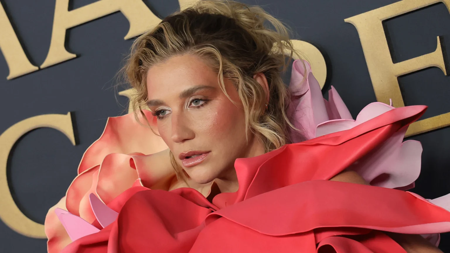 Close-up of Kesha with blonde wavy hair and noticeable makeup, wearing a sculptural garment of layered pink and red fabric resembling large flower petals, against a dark background with gold letters.
