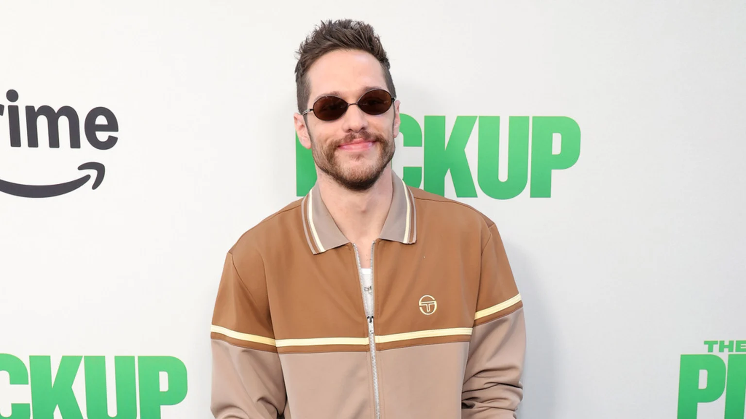 Pete Davidson smiles, wearing oval sunglasses and a brown and beige track jacket with a Sergio Tacchini logo, standing in front of a white wall with "Prime" and "Pickup" logos.