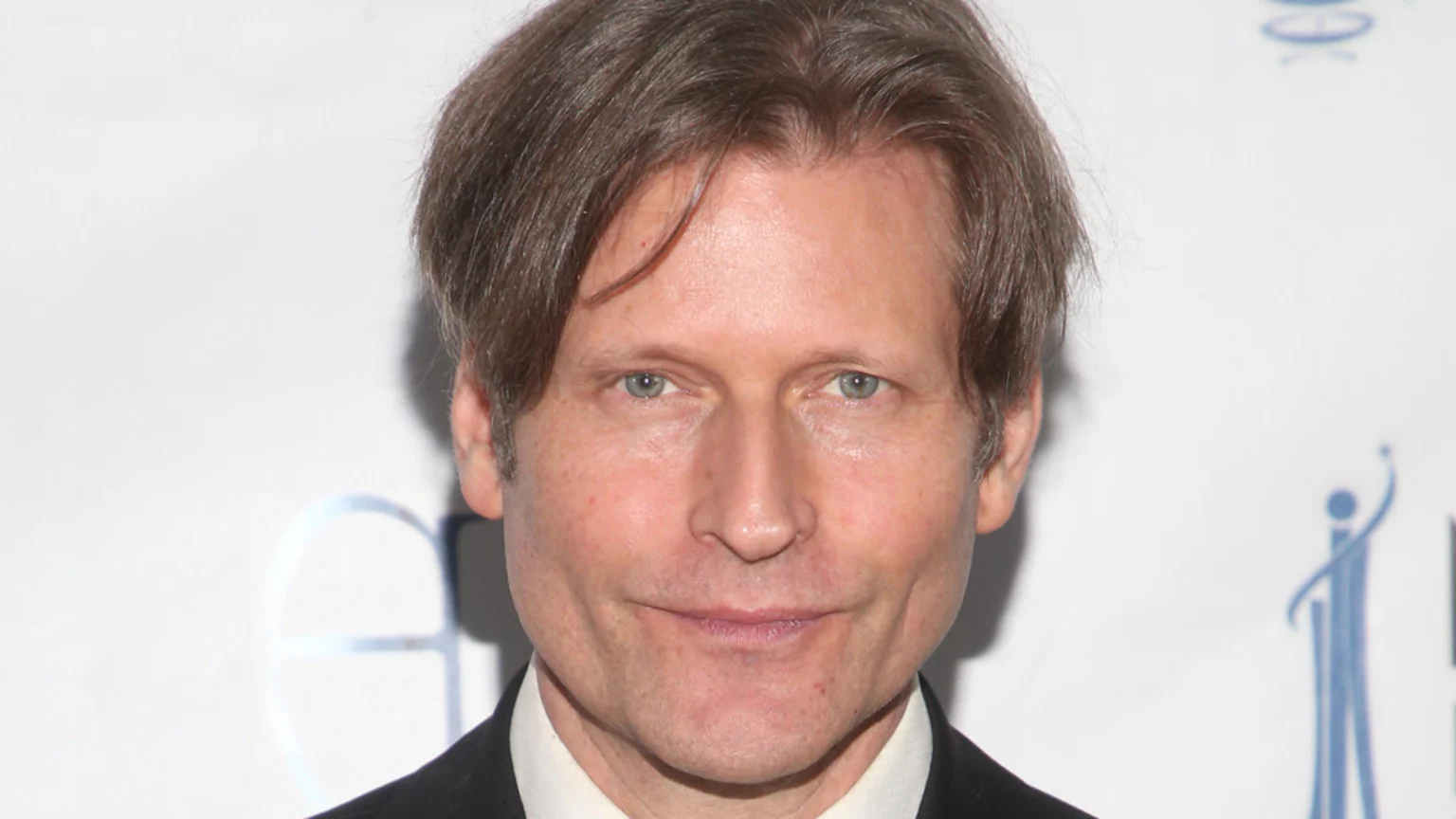 A man with light brown hair and light eyes looks directly at the camera with a neutral expression, wearing a suit jacket and white shirt against a white background.