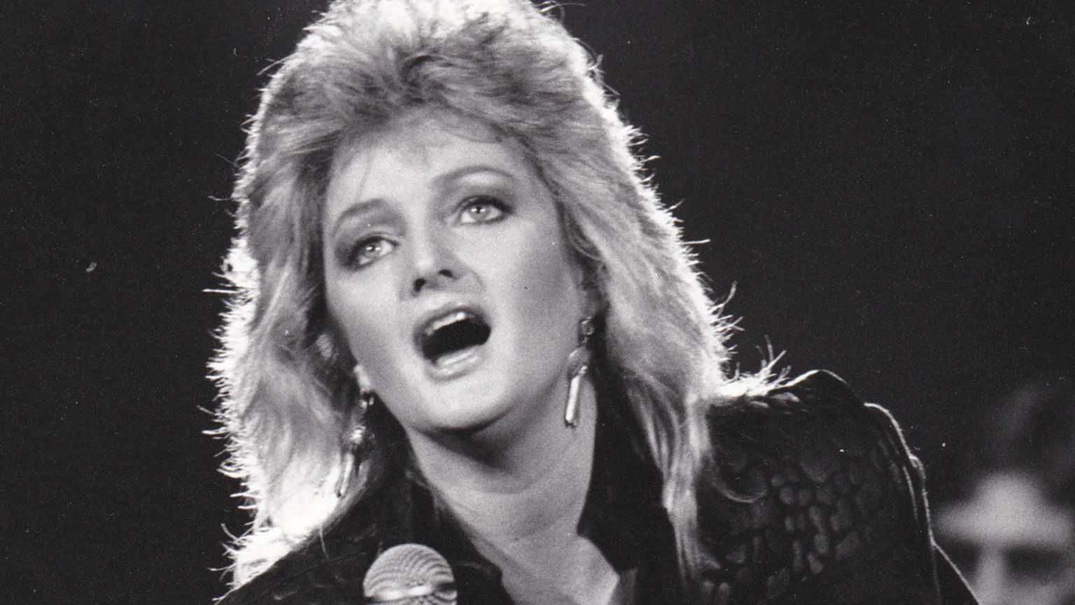 Black and white close-up of a woman with voluminous hair singing into a microphone, her mouth open and looking upwards, with strong backlighting highlighting her hair.