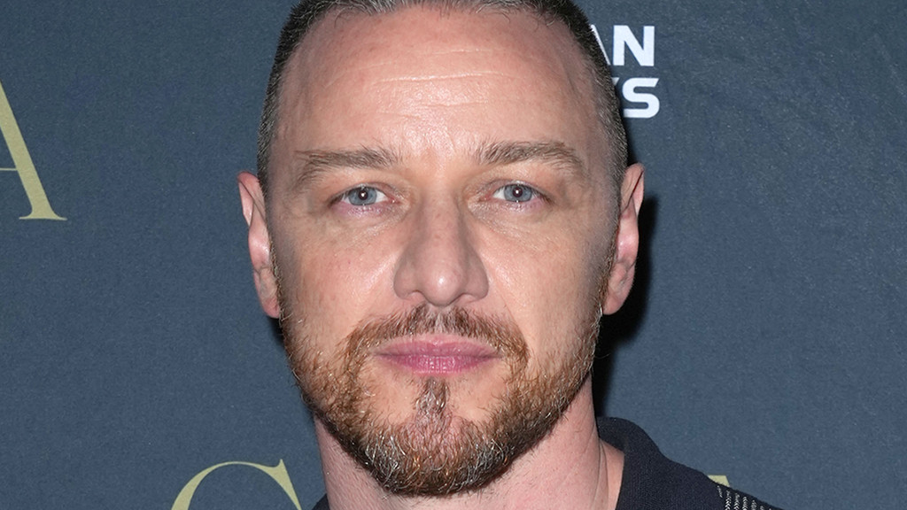 A man with light eyes, a buzzed haircut, and a neatly groomed beard and mustache looks forward against a dark, blurred background.