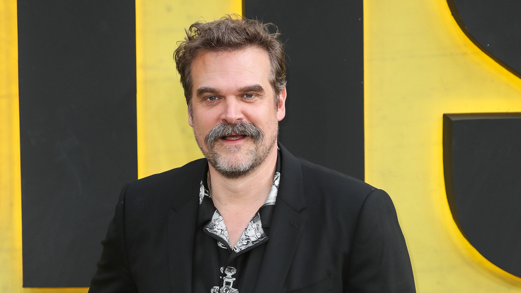 A man with a mustache and beard smiles slightly, wearing a black suit jacket over a dark patterned shirt, against a yellow and black background.