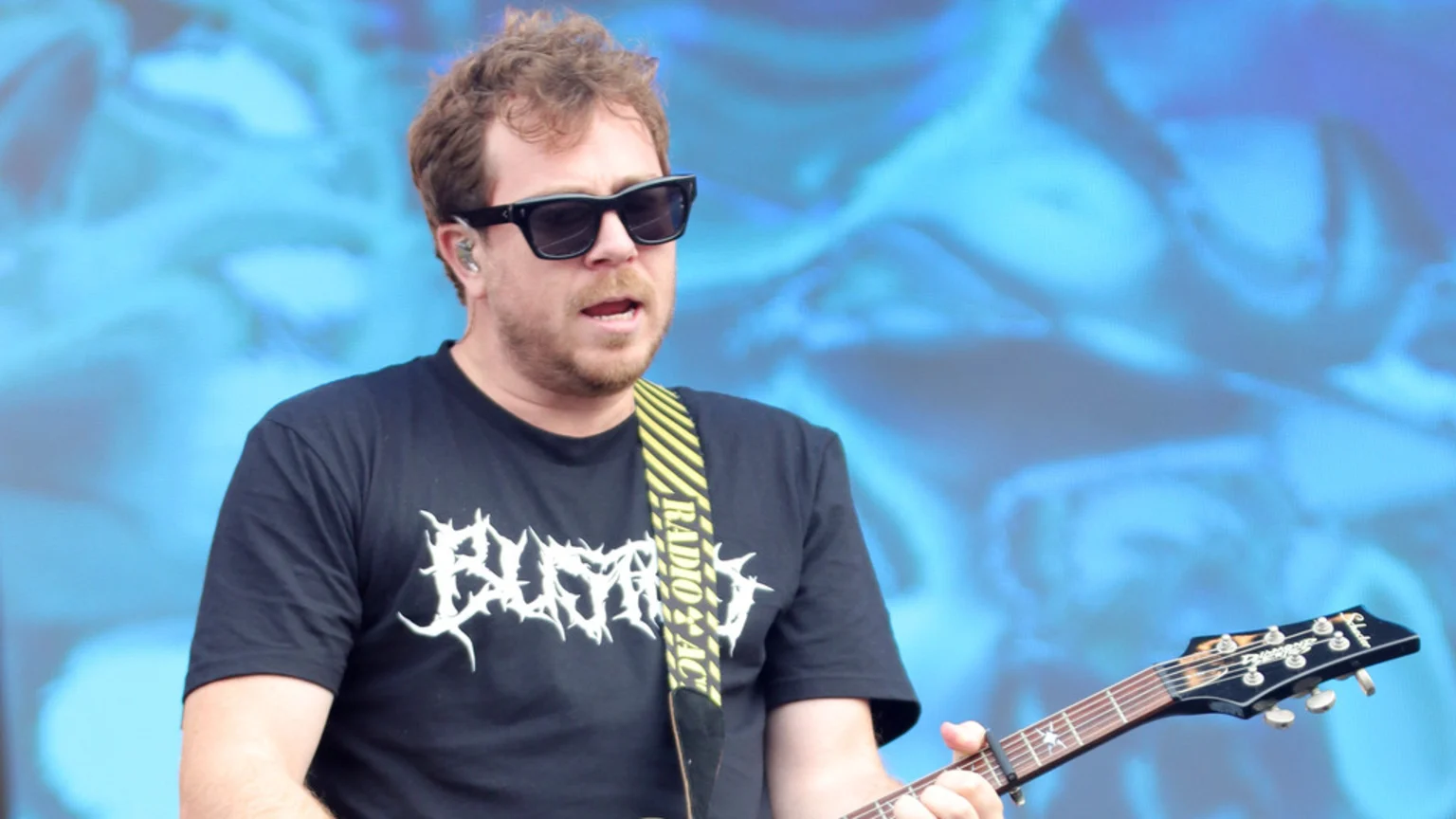 James Bourne plays an Epiphone guitar, wearing sunglasses, a black t-shirt with a jagged white logo, and a "RADIO X" strap, against a blurred blue background.