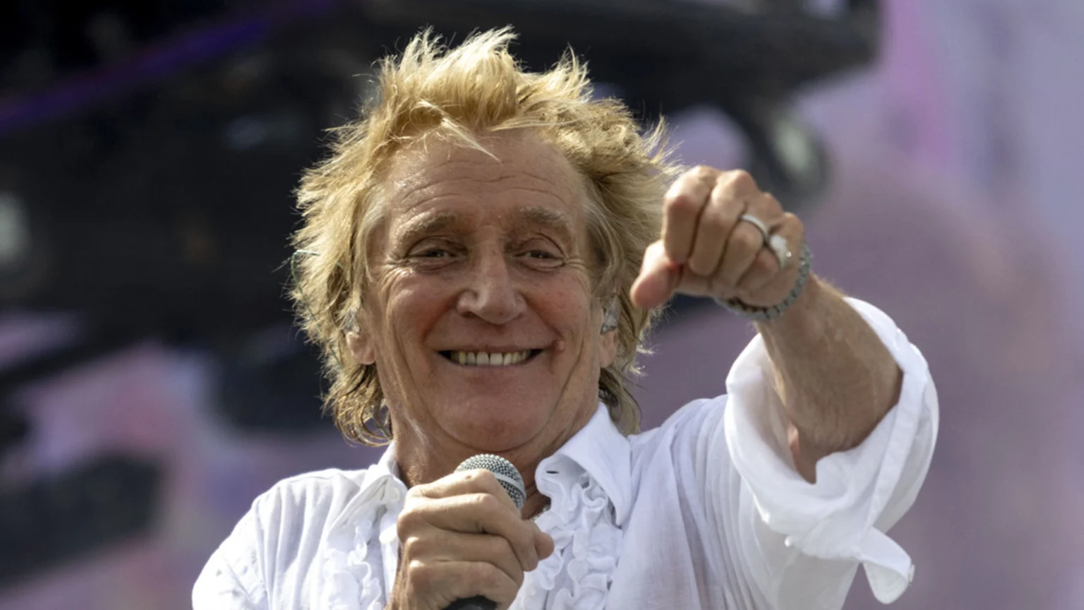 Rod Stewart, with spiky blonde hair and a white ruffled shirt, smiles broadly while holding a microphone and making a thumbs-down gesture with his right hand.